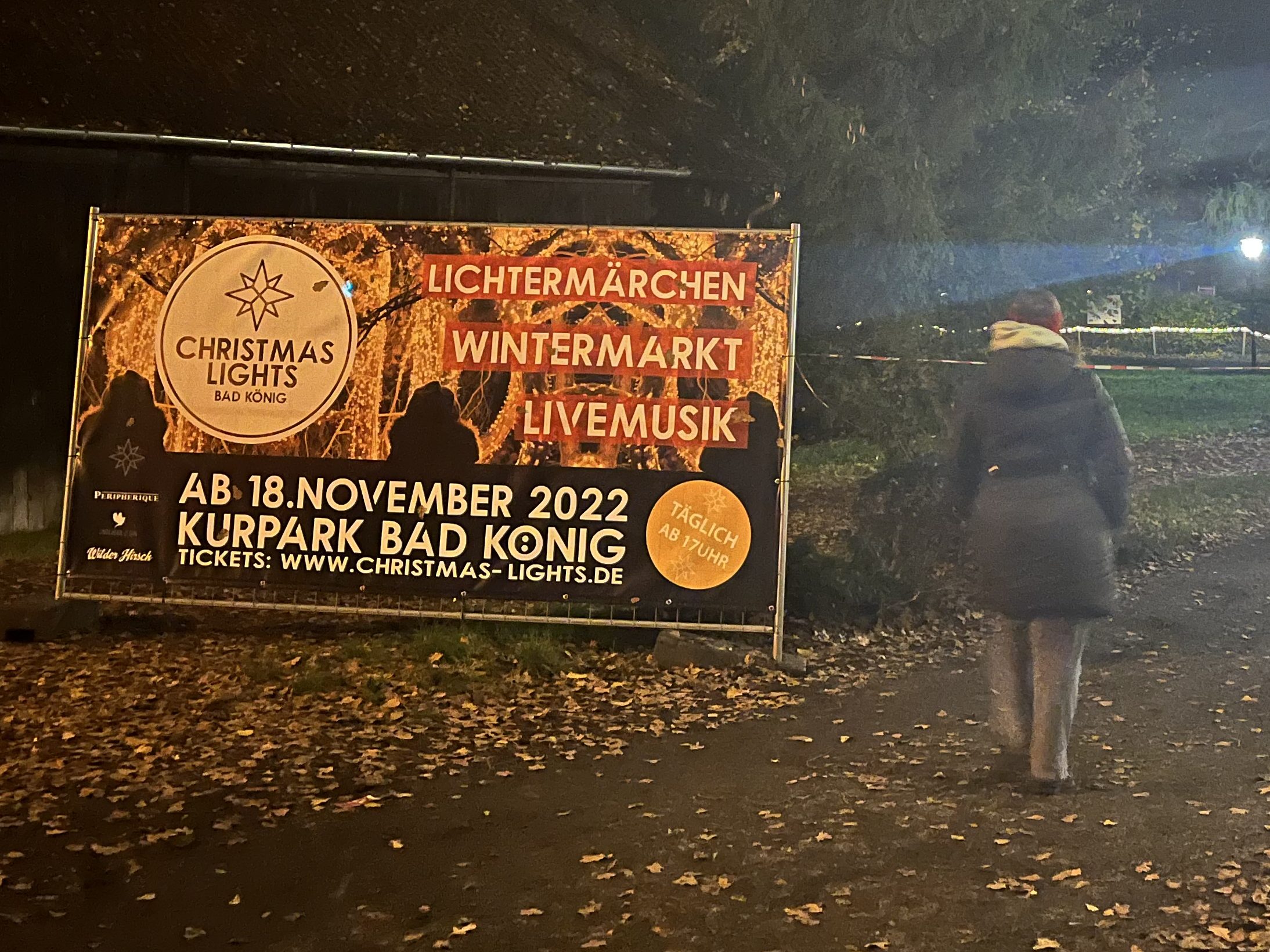 Eine Frau läuft einen Waldweg entlang. An einem Baum hängt eine Lichterkette, neben der Frau steht ein großes Werbeschild mit der Aufschrift Christmas Lights.