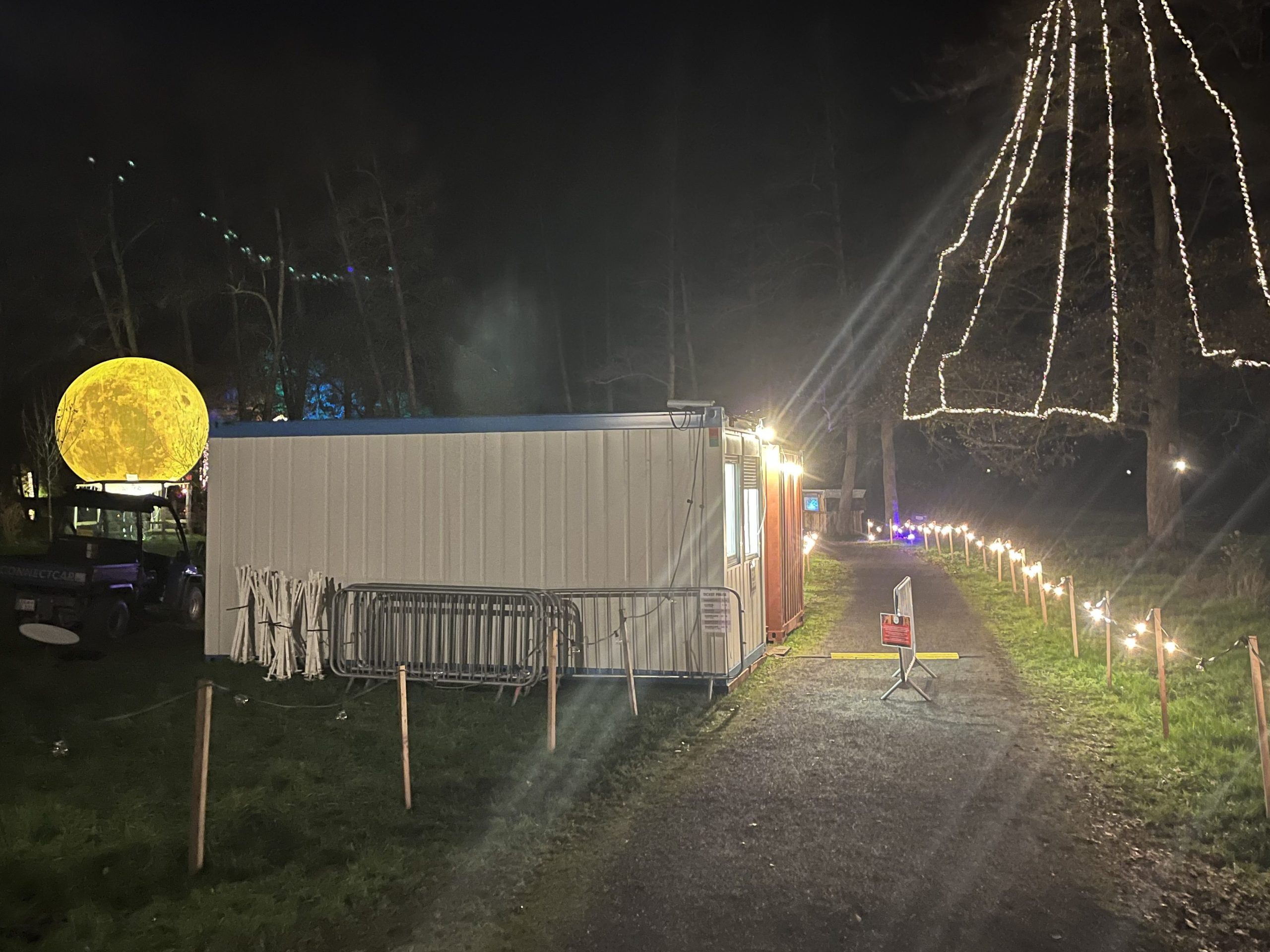 An einem Waldweg steht eine flache weiße Hütte. Im Hintergrund sieht man einen mit Lichterketten geschmückten Baum.