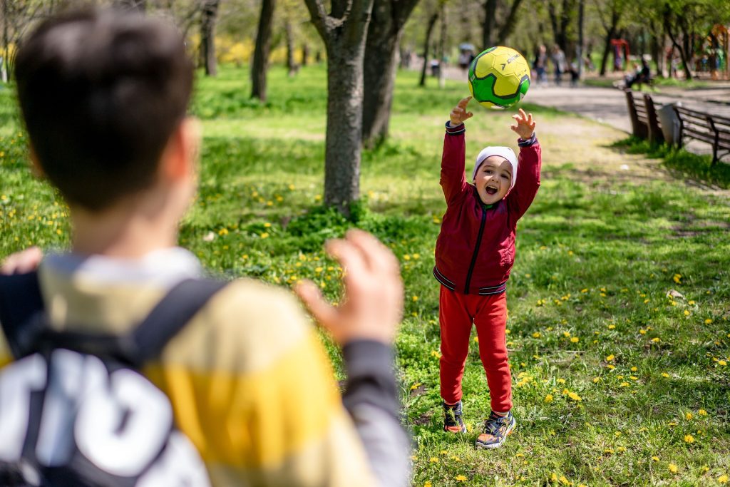 Ein kleines Kind wirft mit offenem Mund einen gelben Ball zu einem älteren Kind.