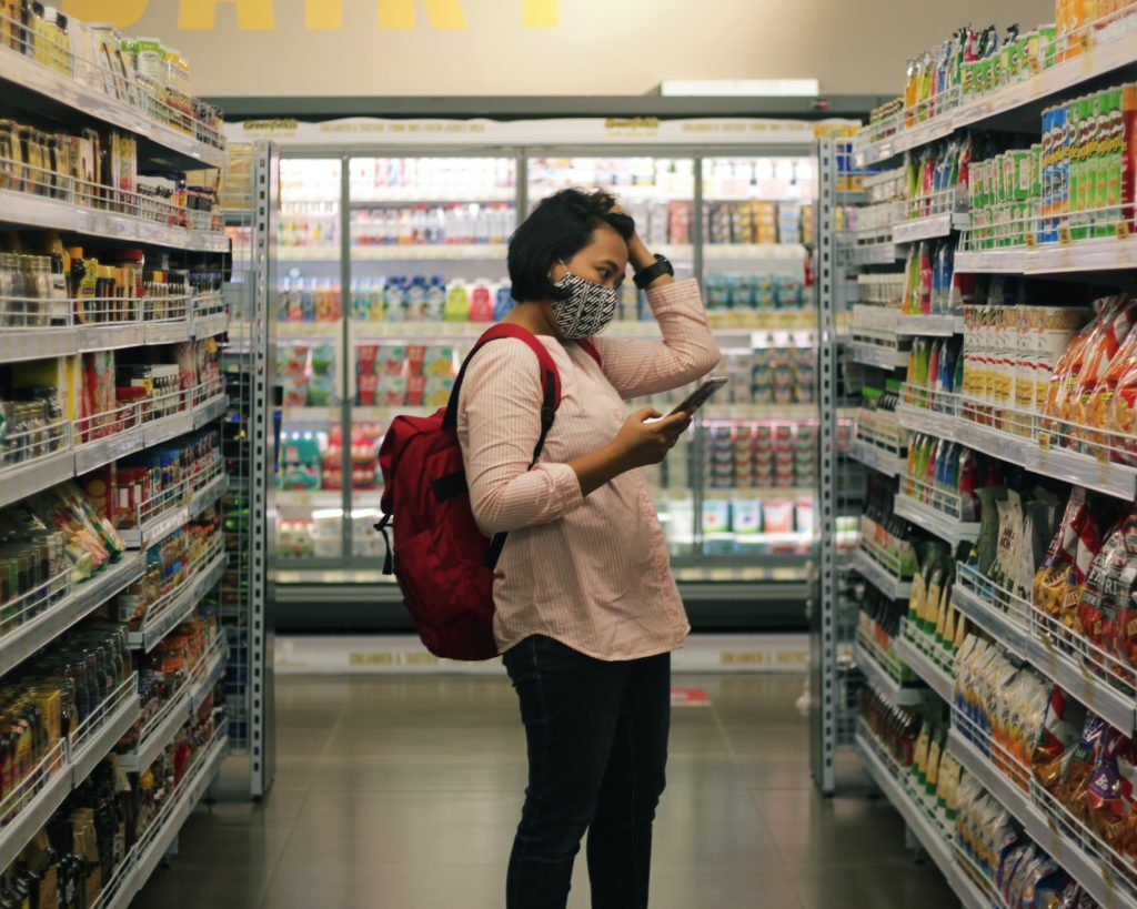 Eine Frau steht mit einem Einkaufszettel vor einem Regal in einem Supermarkt und überlegt.